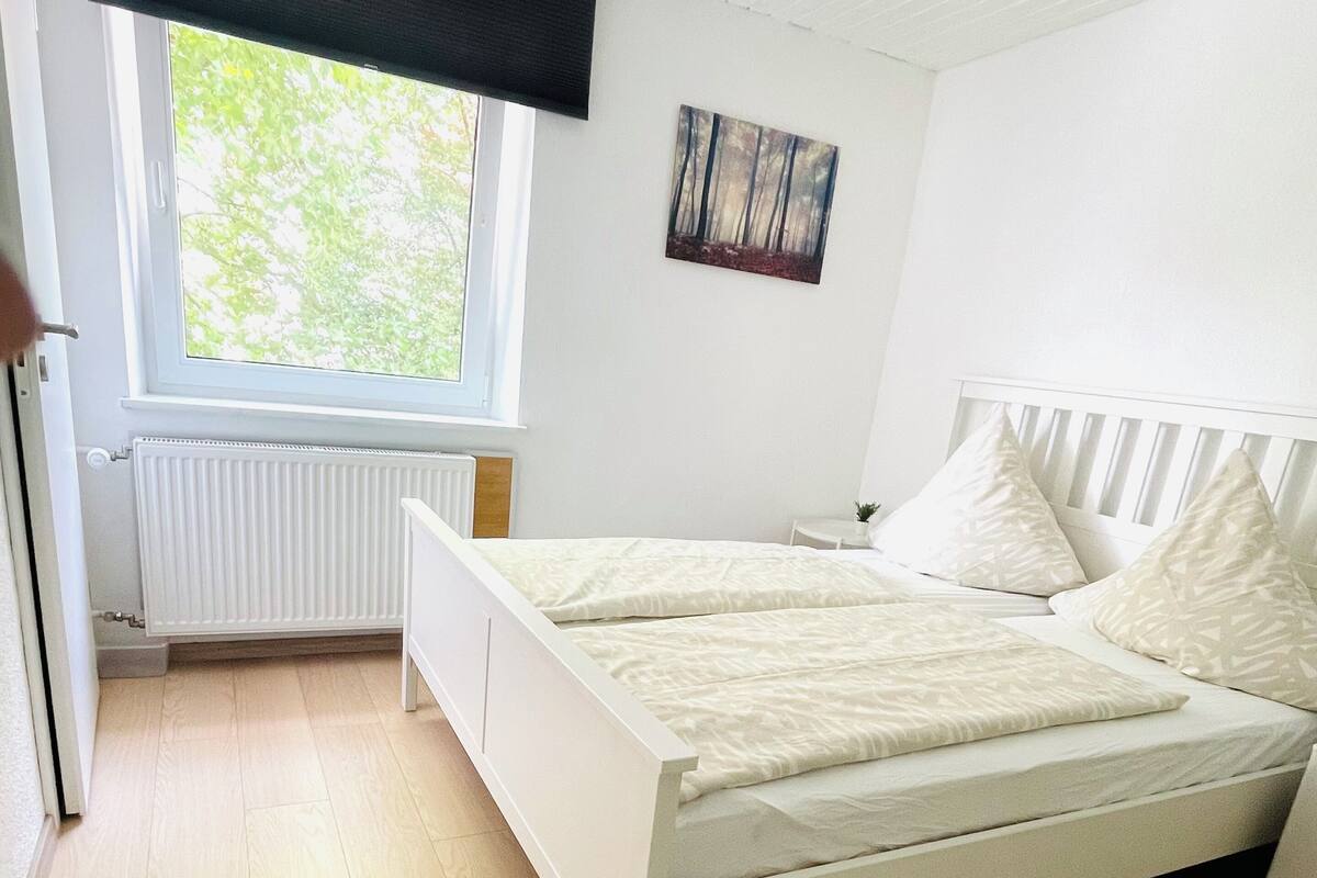 Un dormitorio con una cama blanca, una ventana con vistas a zonas verdes, un radiador y una pequeña mesa blanca.