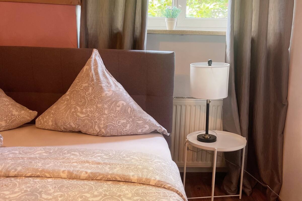 Un dormitorio con una cama grande, una mesita de noche con lámpara y una ventana con cortinas.