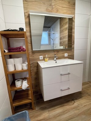 A bathroom with a wooden shelf holding towels, a white sink with a mirror above it, and a wooden floor.