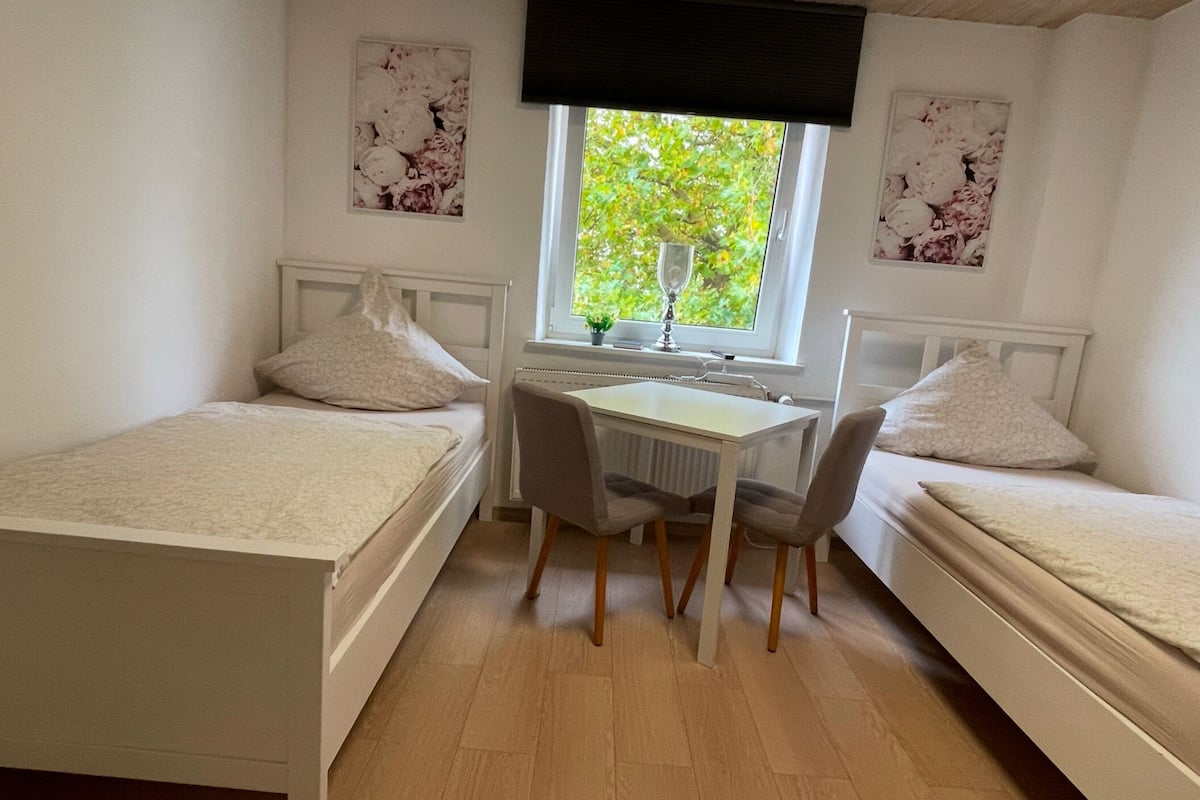 Un dormitorio pequeño y bien iluminado con dos camas individuales, un escritorio con silla, y una ventana con vistas a árboles.
