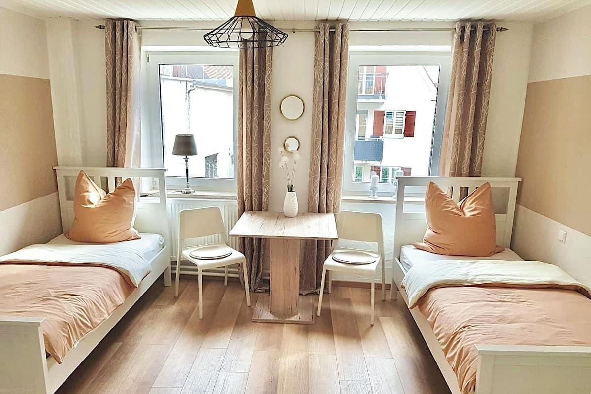 Una habitación con dos camas individuales, una mesita, dos sillas, una lámpara y un ventanal con cortinas.