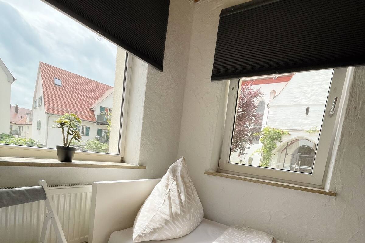 Una habitación pequeña y acogedora con una cama blanca, un banco y una ventana con vistas a edificios.