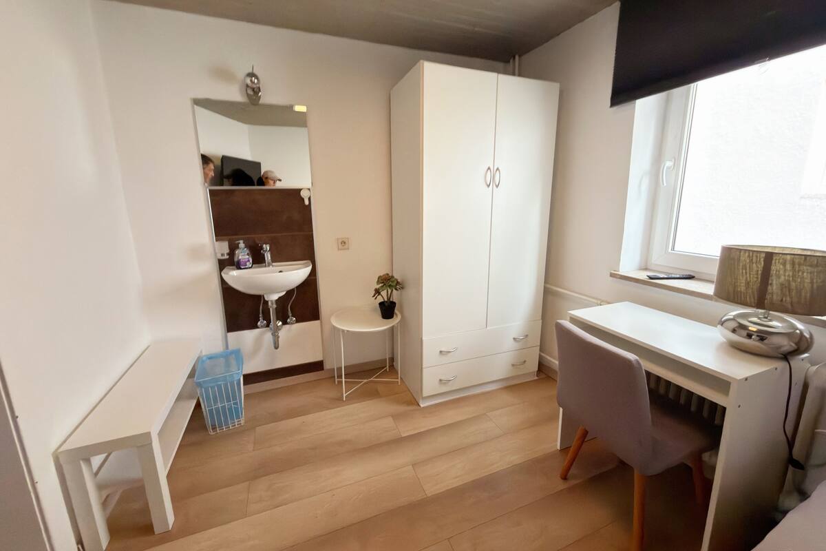 Una habitación pequeña y moderna con un escritorio blanco, un armario blanco, una mesita redonda con una planta, un lavamanos blanco y una ventana con cortina.