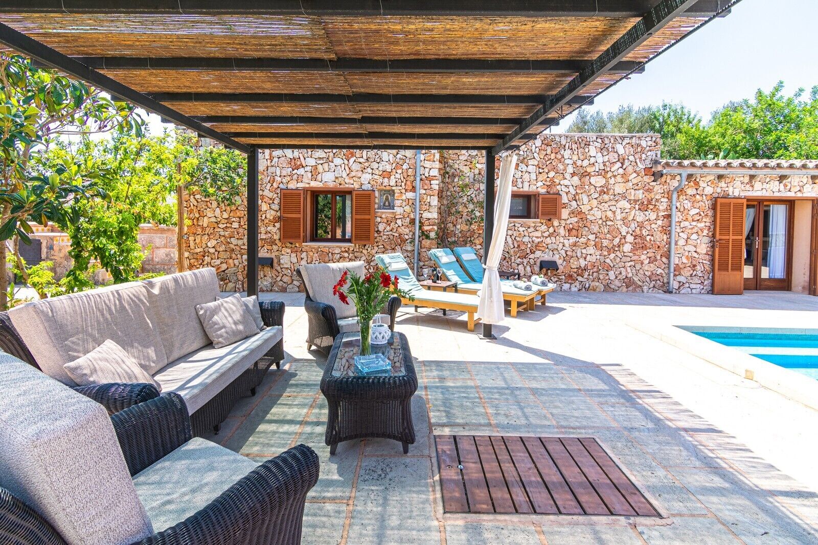 A covered outdoor seating area with wicker furniture, a stone wall, and a pool.