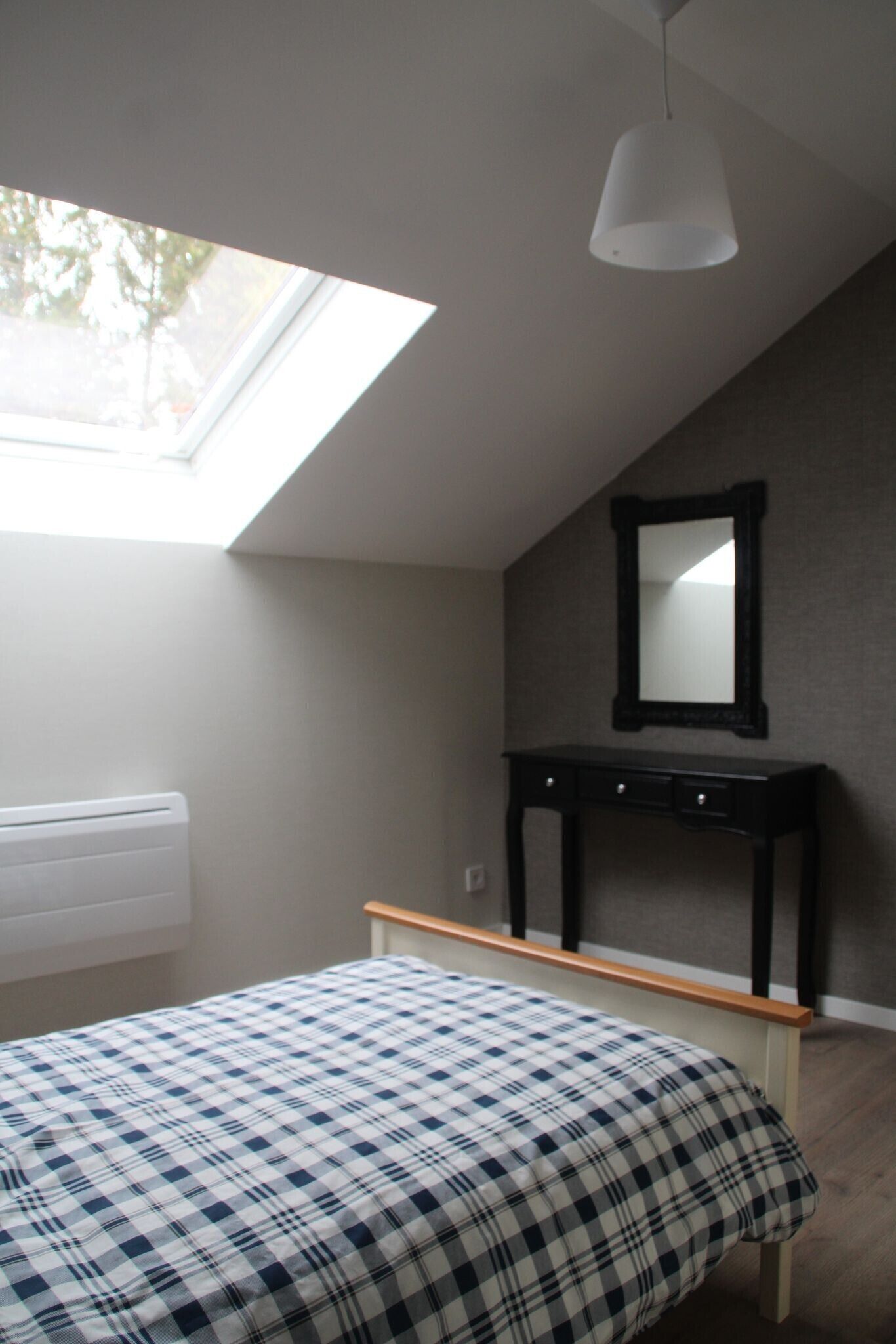 A bedroom with a skylight, a bed with a checkered bedspread, a bedside table, a mirror, and a radiator.