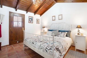 A bedroom with a wooden ceiling, a white bed with a floral bedspread, two bedside tables with lamps, and a wooden door.