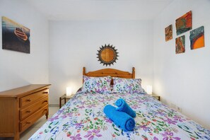 A bedroom with a wooden bed, floral bedding, a wooden dresser, and a sun-shaped wall decoration.