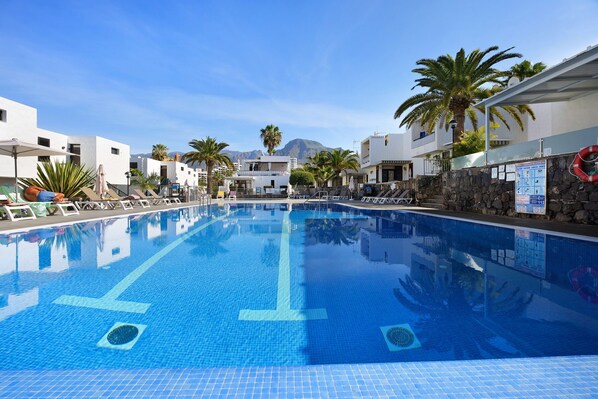 A swimming pool with clear blue water, surrounded by white buildings and palm trees.