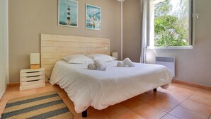 Un dormitorio con cabecera de madera, cama con ropa blanca, dos carteles en la pared, una ventana con vista a la vegetación y una mesita de noche con lámpara.