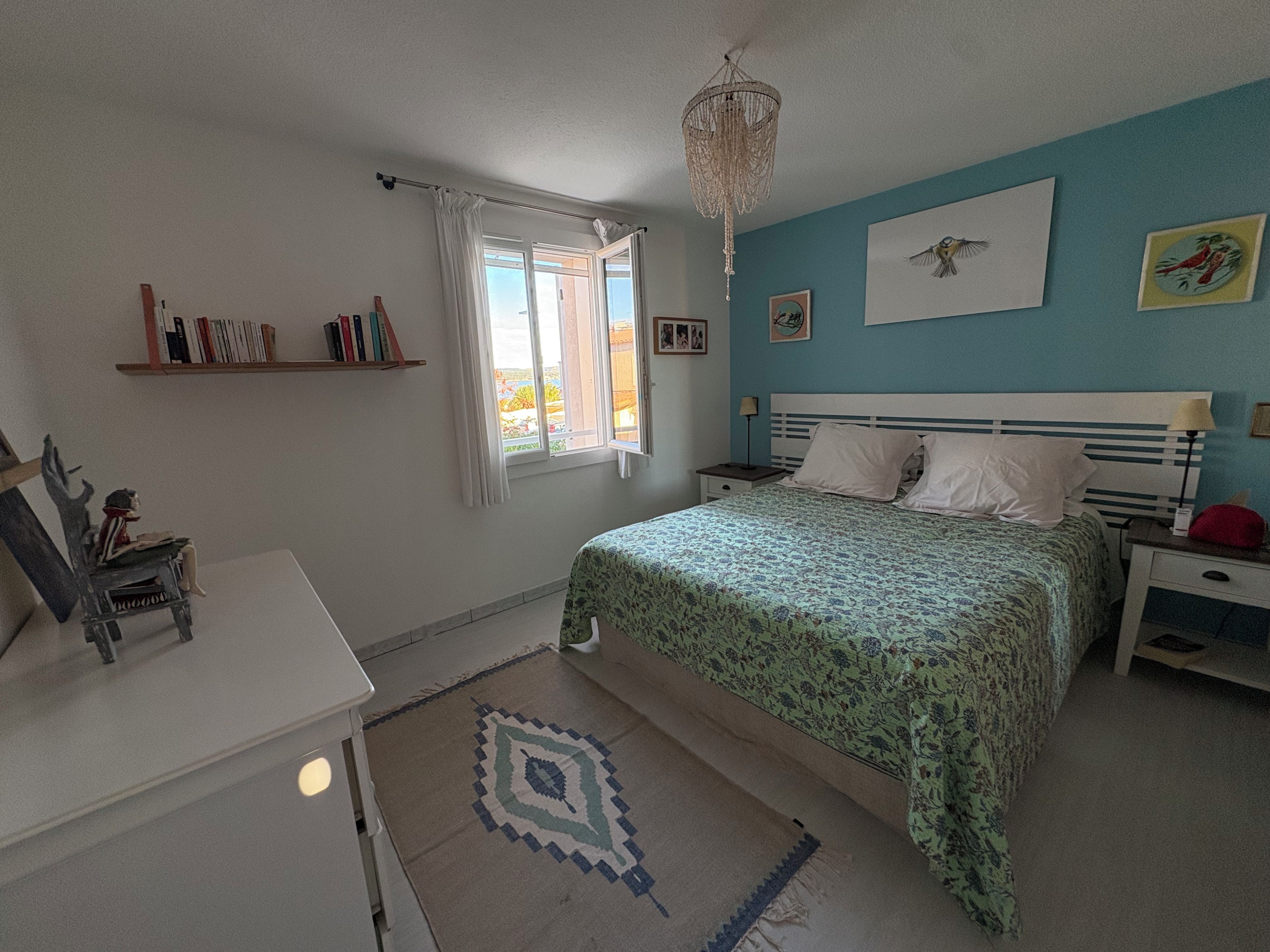 A bedroom with a bed, a nightstand, a desk, a chair, a window with curtains, a chandelier, and wall art.