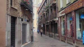 Uma rua estreita com edif ícios históricos, um campanário de igreja ao fundo e lojas fechadas.