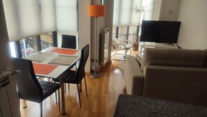 Uma sala de jantar com uma mesa, cadeiras e um abajur. Há um sofá e uma TV na sala de estar.