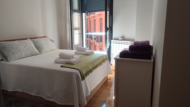 Quarto de hotel com cama, janela com vista para um edifício e uma mesa pequena com toalhas.