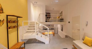 Espaço compacto com escada conectando dois níveis, cozinha compacta, área de jantar e sofá com almofada amarela.