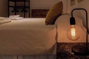 Cama bem arrumada com lençóis brancos e um único travesseiro amarelo. Uma luminária de mesa moderna com cúpula de vidro transparente está sobre a mesa de cabeceira.
