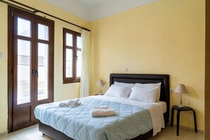A hotel room with a bed, two bedside lamps, a window with curtains, and a door with frosted glass panels. - George's Poet Maisonette (Rethymno)