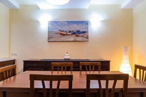 A dining area with a wooden table and chairs, a painting of boats on the wall, and a lamp on the right. - George's Poet Maisonette (Rethymno)
