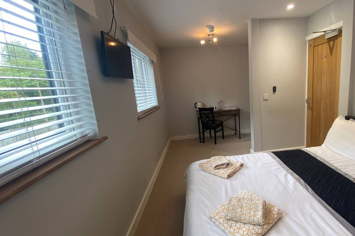 A bedroom with a bed, a desk, and a chair. There is a window with blinds, a ceiling fan, and a door.