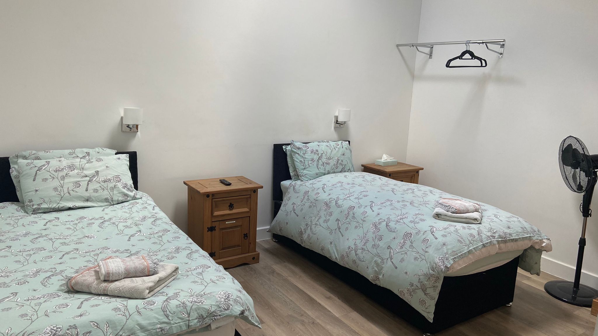 A bedroom with two single beds, wooden nightstands, a bedside table, a hanging clothes rack, and a standing fan.