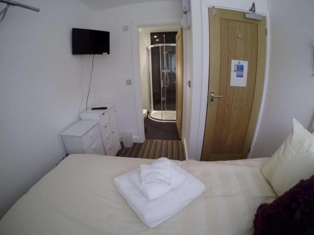 Une chambre d’hôtel avec un lit, un téléviseur fixé au mur, une porte en bois et un espace douche visible à travers une porte ouverte.