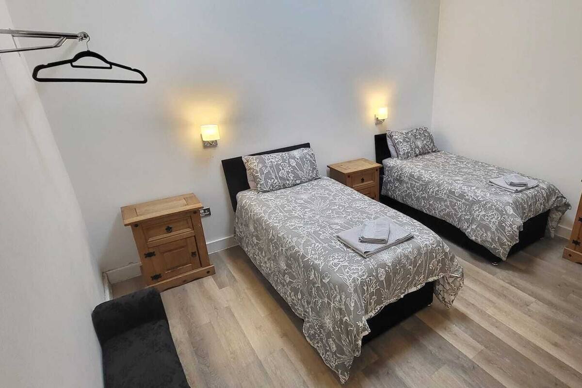 A bedroom with two beds, bedside tables, a hanging clothes rack, a floor fan, and a wall-mounted TV.