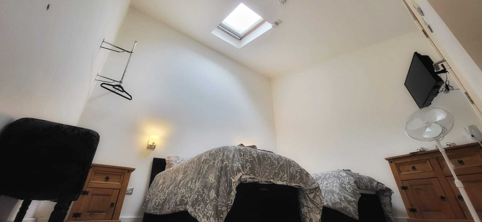 A bedroom with a skylight, a bed with floral bedding, a wooden dresser, and a standing fan.