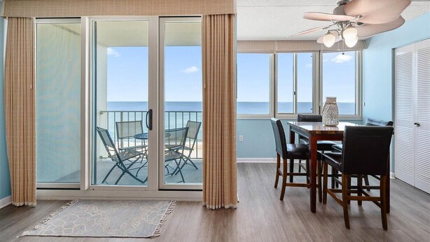 A room with a sliding glass door leading to a balcony with a view of the ocean. There is a dining table with chairs and a ceiling fan. - Quay-3 Bedroom Ocean Front Condo (Ocean City)