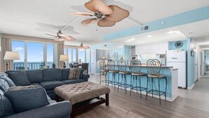 A spacious living area with a sofa, ottoman, and ceiling fans, leading to a kitchen with bar stools and a refrigerator. - Quay-3 Bedroom Ocean Front Condo (Ocean City)