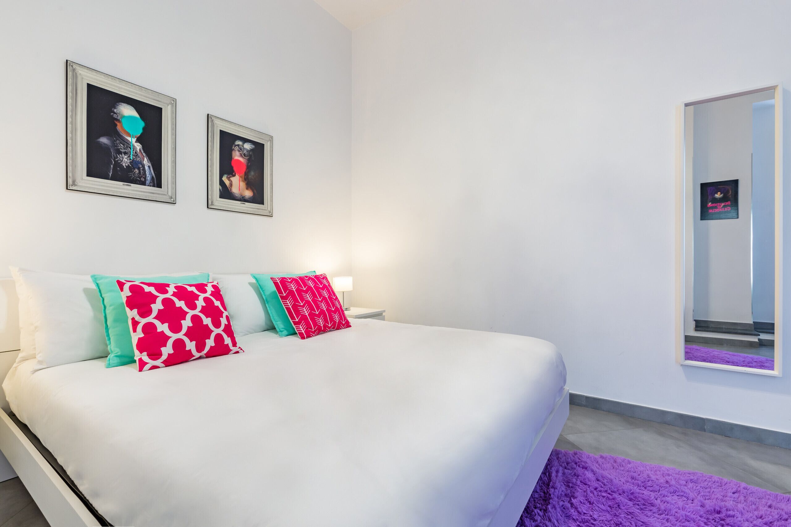 Ein modernes Schlafzimmer mit einem großen Bett, weißer Bettwäsche und farbenfrohen Kissen. Zwei gerahmte Kunstwerke sind an der Wand angebracht und ein Spiegel spiegelt den Raum wider.