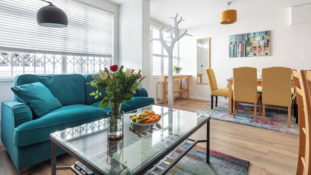 A modern living room with a teal sofa, a glass coffee table with fruit, and a dining area with yellow chairs.