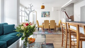 A modern living room with a teal sofa, a glass coffee table with fruit, and a dining area with yellow chairs and a wooden table.