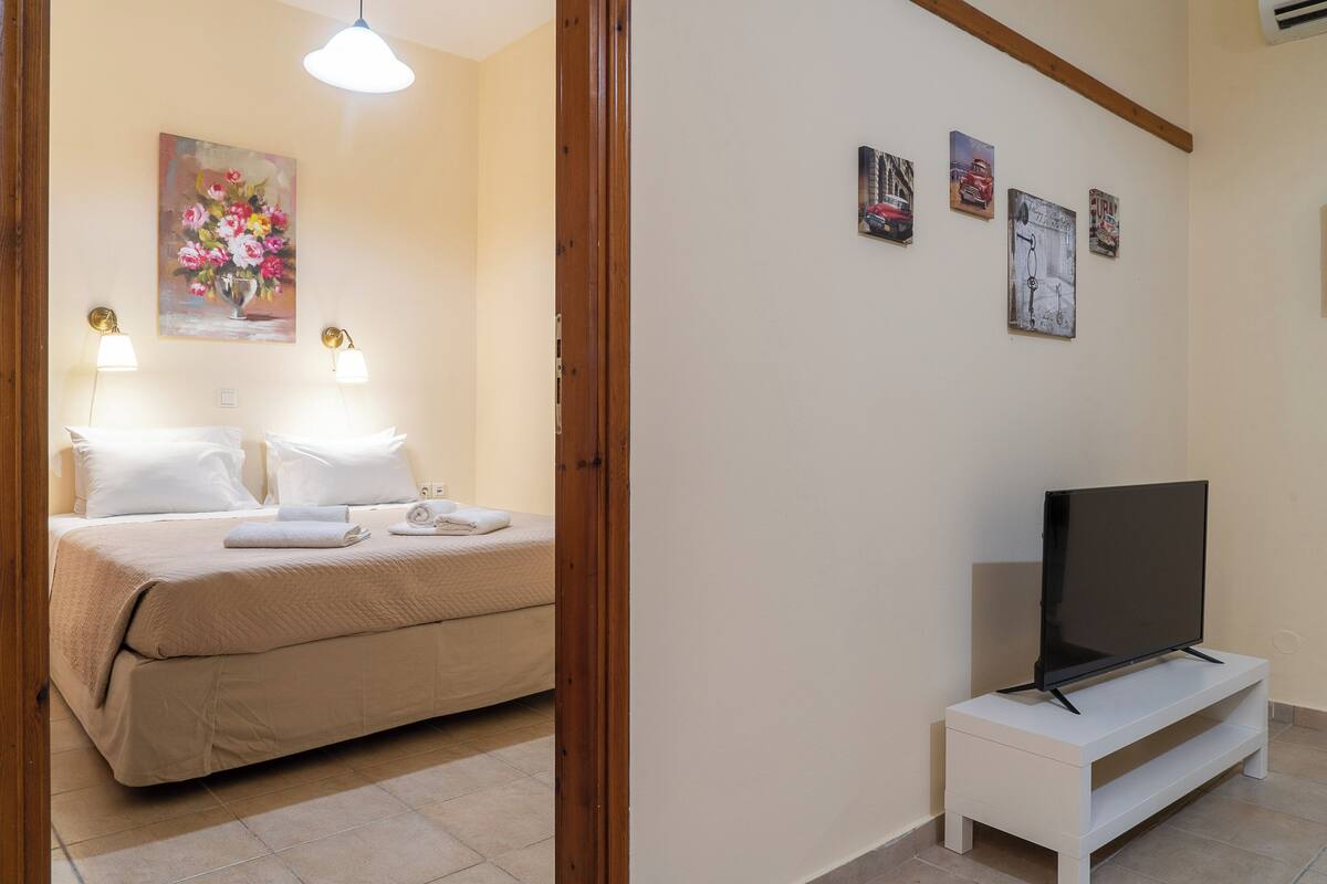 A hotel room with a bed, a TV on a white stand, and wall art including a floral painting and three small pictures.