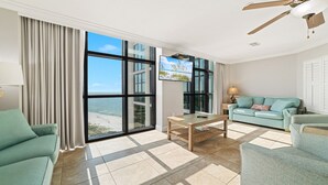 A spacious living room with a large window offering a view of the beach, a television, and a ceiling fan. - Oasis, formerly Phoenix West II 4BR!!  New Adult Pool,  Lazy River/Slide!! (Orange Beach)