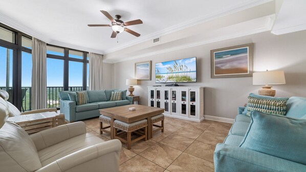 A spacious living room with a large window, a ceiling fan, a flat-screen TV, and a dining area. - Oasis, formerly Phoenix West II 4BR!!  New Adult Pool,  Lazy River/Slide!! (Orange Beach)