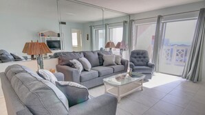 A modern living room with a grey sofa, armchairs, and a glass coffee table. A TV is mounted on the wall, and there is a balcony with curtains.