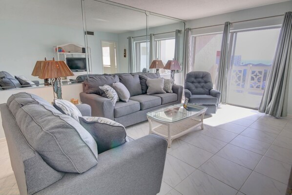 Ein modernes Wohnzimmer mit einem grauen Sofa, Sesseln und einem Glastisch. Ein Fernseher ist an der Wand montiert, und es gibt einen Balkon mit Vorhängen.