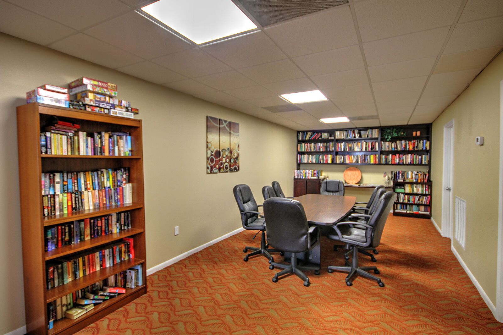 A library with a large wooden bookshelf, a round table with chairs, and a patterned carpet.