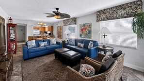 A living room with a blue sofa, a black ottoman, a wicker chair, and a ceiling fan. - 110 El Matador 2 Bedroom Condo by RedAwning (Fort Walton Beach)