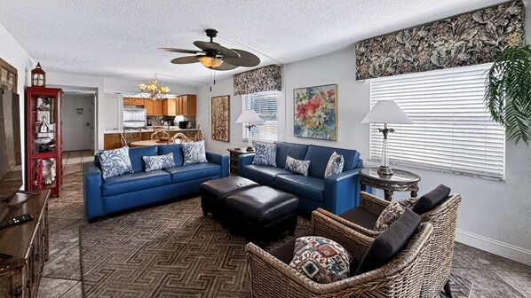 A living room with a blue sofa, a black ottoman, a wicker chair, and a ceiling fan.
