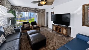Una sala de estar moderna con sofá, reposapiés y sillones, un televisor y vistas a un edificio y palmeras.