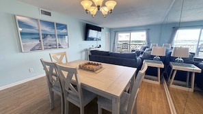 Ett rum med ett matbord i trä, en soffa, en tv som är monterad på väggen och utsikt över havet genom stora fönster.