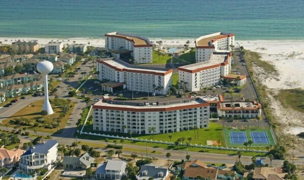 Un complejo hotelero frente al mar con varias construcciones, una torre blanca grande y una playa al fondo.