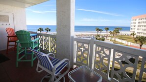A balcony with chairs and a table, overlooking a beach with palm trees and a building in the distance.