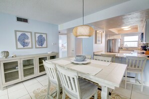 A dining area with a white table and chairs, a hanging light fixture, and a kitchen in the background. - 358 El Matador 2 Bedroom Condo by RedAwning (Fort Walton Beach)