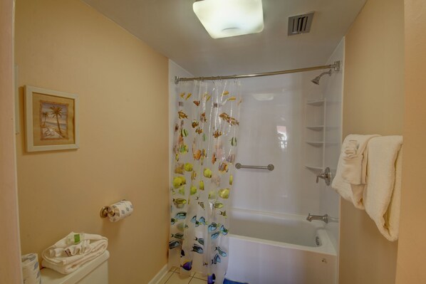 Une salle de bain avec un rideau de douche orné de motifs de poissons, une baignoire, des toilettes avec une serviette à proximité, et un tableau encadré accroché au mur.