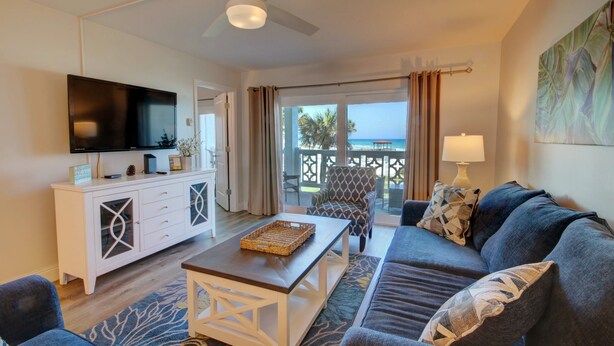 A modern living room with a flat-screen TV, a white cabinet, a blue sofa, a patterned armchair, and a view of the ocean through large windows. - 427 El Matador 2 Bedroom Condo by RedAwning (Fort Walton Beach)