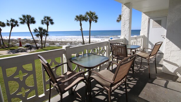Un balcon avec une table et des chaises en verre, donnant sur une plage bordée de palmiers et offrant une vue sur un ciel dégagé.