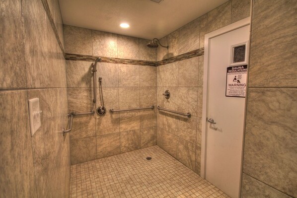 Pequeño espacio de ducha con azulejos, barras de apoyo y un cartel con las normas de la sauna.