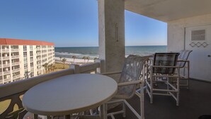 A balcony with a table and chairs, overlooking a beach and a hotel building.