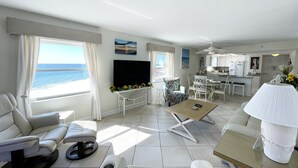 A modern living room with a sofa, a coffee table, and a TV. There is a view of the ocean through large windows.